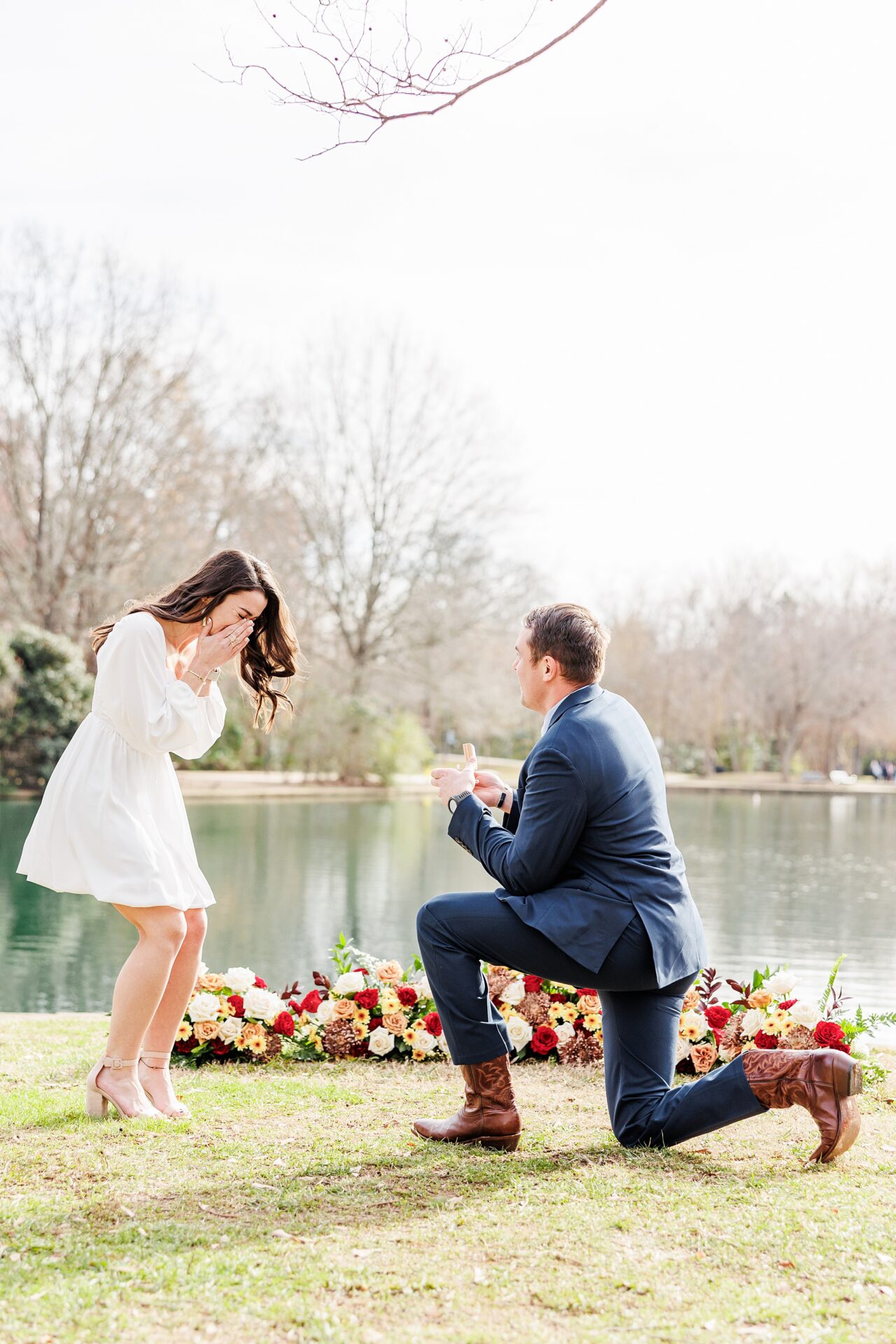 David on one knee proposing to Amanda beside the lake at Freedom Park, her hands over her mouth in joyful surprise. Charlotte Surprise Proposal captured by a Surprise Proposal Photos, featuring Proposal outfit ideas and Floral Grounded Arch.